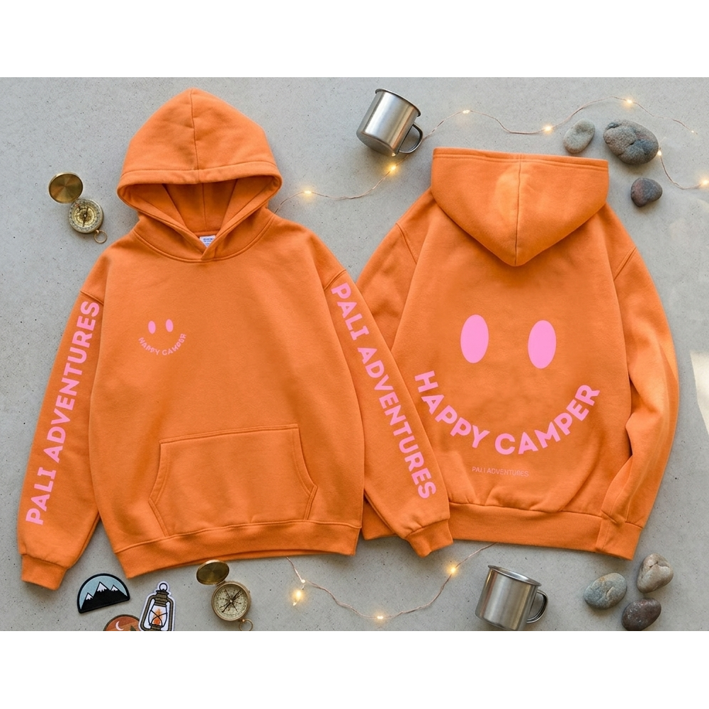Orange hoodie with 'Happy Camper' text and 'Palli Adventures' branding on a gray surface with decorative elements.